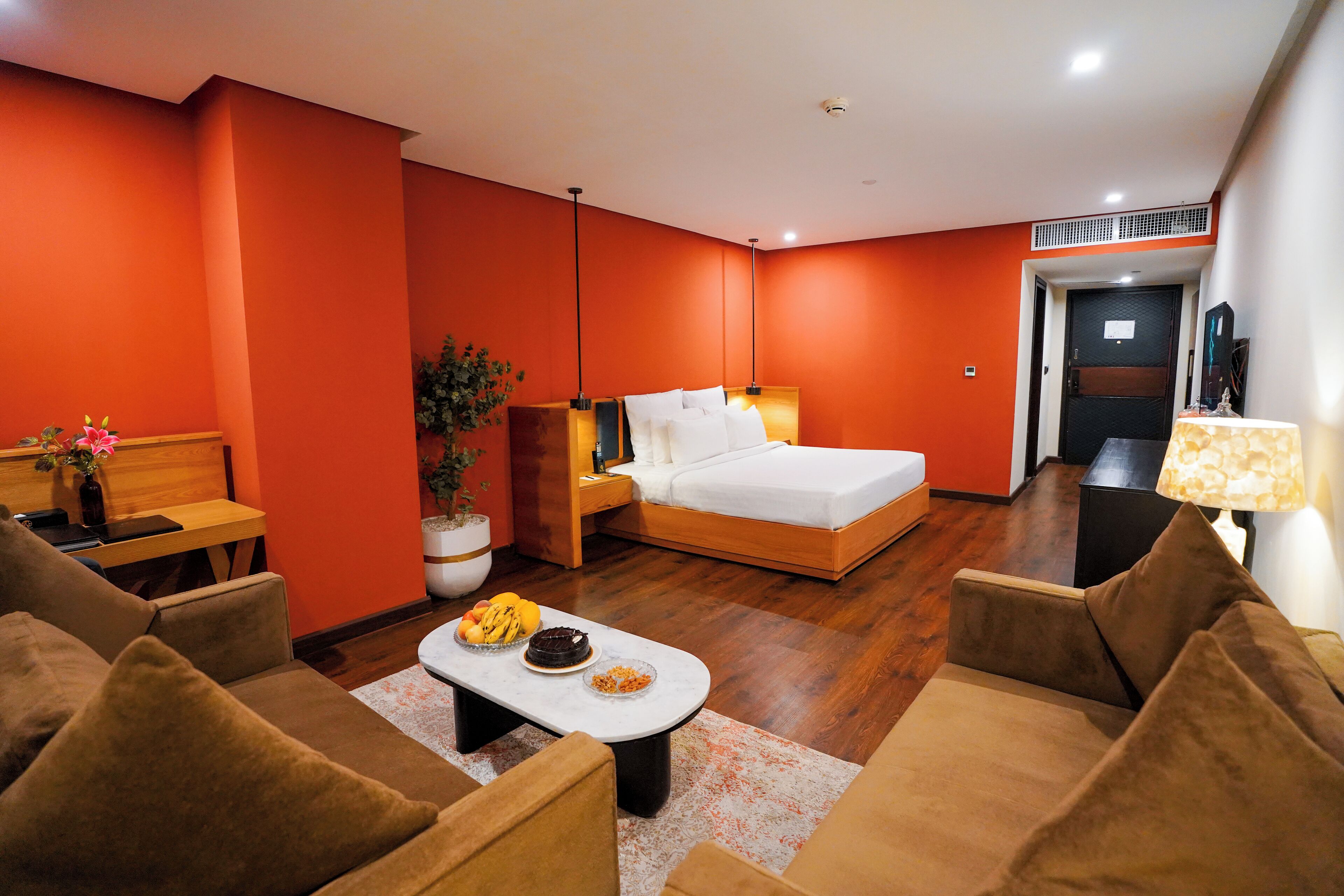 Habitación de hotel con una cama, dos sofás, una mesa de centro con frutas, una planta y un escritorio con una silla.