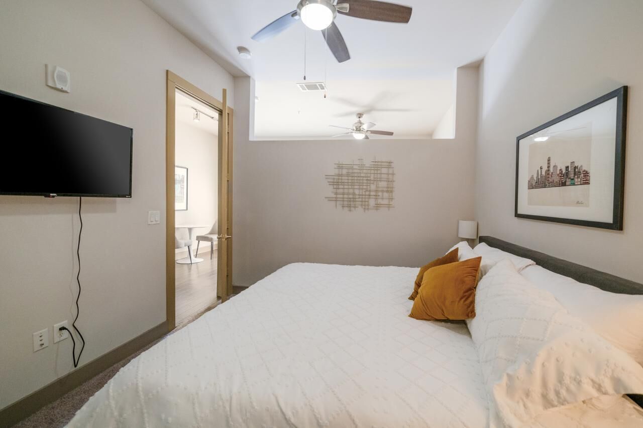 Une chambre à coucher avec un lit, un ventilateur de plafond, une télévision et un tableau représentant une vue de la ville, encadré.