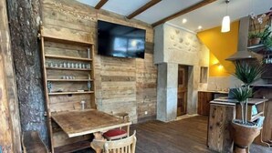 Un intérieur rustique avec des poutres apparentes, un banc en bois, une table avec des chaises, un téléviseur à écran plat fixé au mur et une cheminée en pierre.