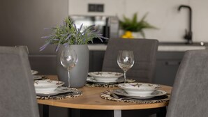 Una mesa de comedor dispuesta con platos blancos, cubiertos y dos copas de vino. Un florero gris con flores moradas está sobre la mesa.