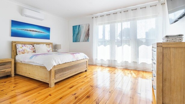 Een slaapkamer met een houten bed, een nachtkastje, een dressoir en een groot raam met dunne gordijnen.