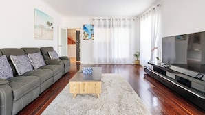 Een moderne woonkamer met een grijze bank, een houten salontafel, een flatscreen-televisie en een groot raam met lichte gordijnen.