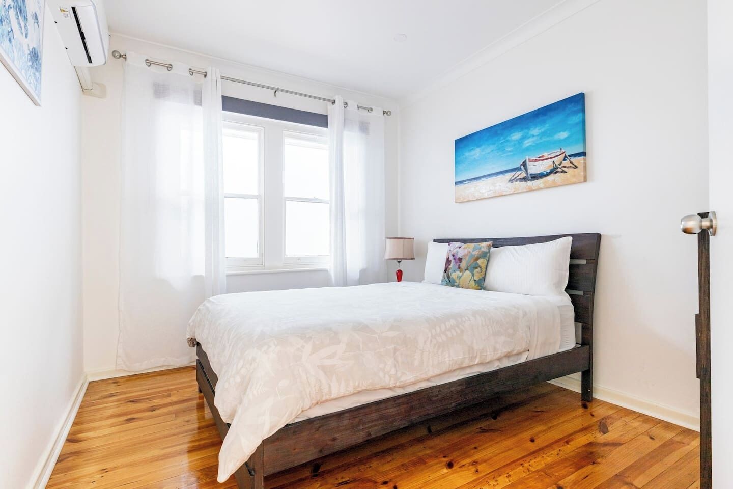 Une chambre à coucher avec un lit en bois, du linge de lit blanc, un tableau représentant une scène de plage et une fenêtre ornée de rideaux légers.