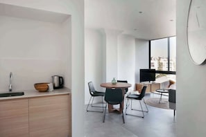 A modern kitchen with a sink, kettle, and wooden bowl, a dining area with a round table and chairs, and a large window with a view of buildings. - The Bright Oasis - Design 1 BR unit in Poble Nou (Barcelona)