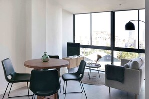 A modern living room with a round wooden table, black chairs, a sofa, and a large window with a city view. - The Bright Oasis - Design 1 BR unit in Poble Nou (Barcelona)
