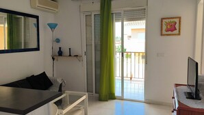 A room with a bed, a desk, a chair, a TV on a wooden table, a window with a green curtain, and a door leading to a balcony.