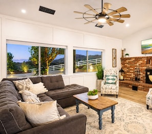 A living room with a sectional sofa, a coffee table, a patterned armchair, a fireplace, and large windows with a view of a landscape. - 25% Off July 20-23! Perfect 4 Families Pool, Spa* (Temecula)
