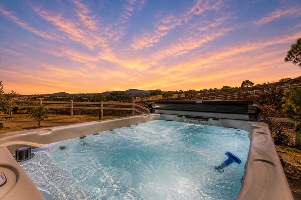 Un jacuzzi con terraza de madera, rodeado por una valla y árboles, con un atardecer al fondo.