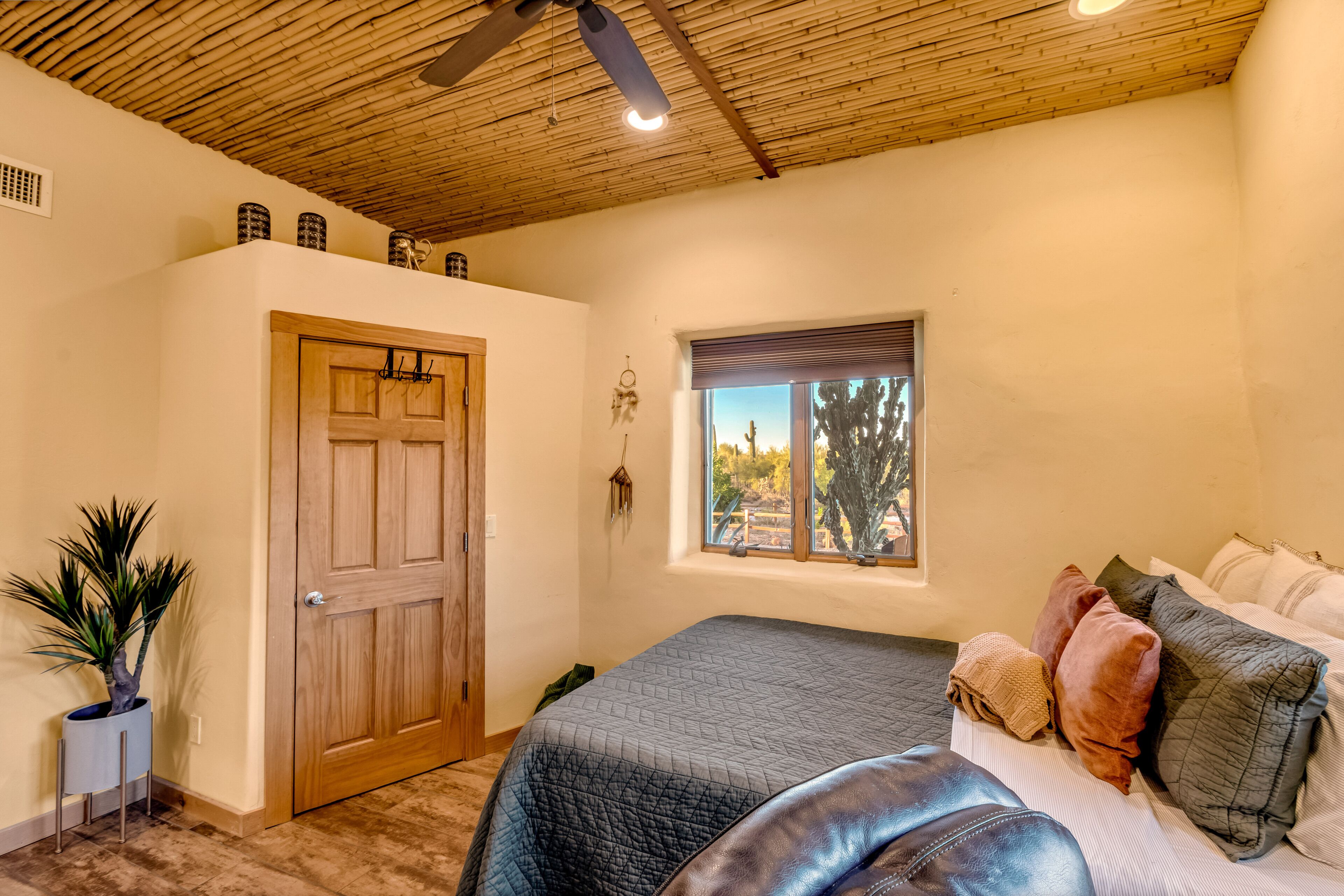 Une chambre avec un plafond en bois, un grand lit avec de nombreux oreillers, une fenêtre donnant sur des arbres et une porte à côté de laquelle se trouve une plante.