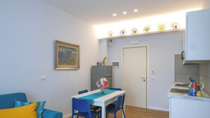 Ein gemütlicher Wohnraum mit Esstisch, blauen Stühlen, einem Sofa mit gelben Kissen, einem Bild an der Wand und einer Küchenzeile mit Mikrowelle und Kaffeemaschine.