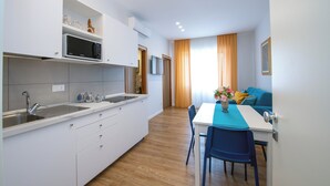 Eine moderne Küche mit weißen Schränken, einer Edelstahlspüle und einer Mikrowelle. Es gibt einen Essbereich mit Tisch und Stühlen, und im Hintergrund steht ein blaues Sofa.