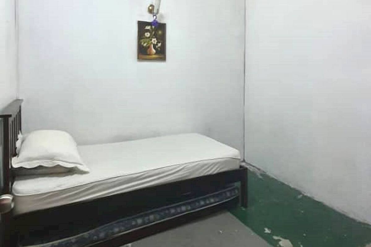 A small, sparsely furnished room with a single bed, a small table, and a wall-mounted light fixture.