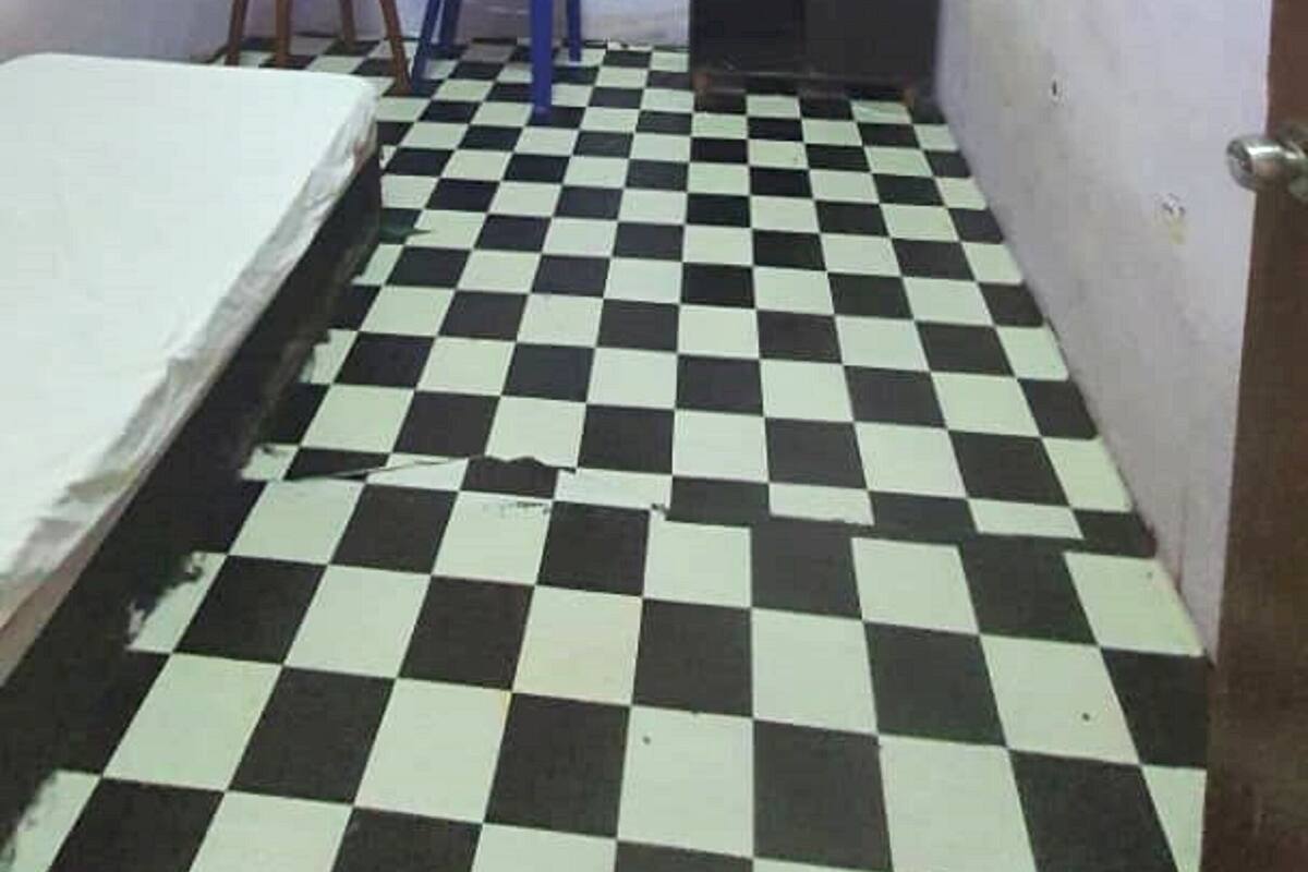 A small, narrow room with a checkered floor, a bed, a desk, a chair, and a ceiling fan.
