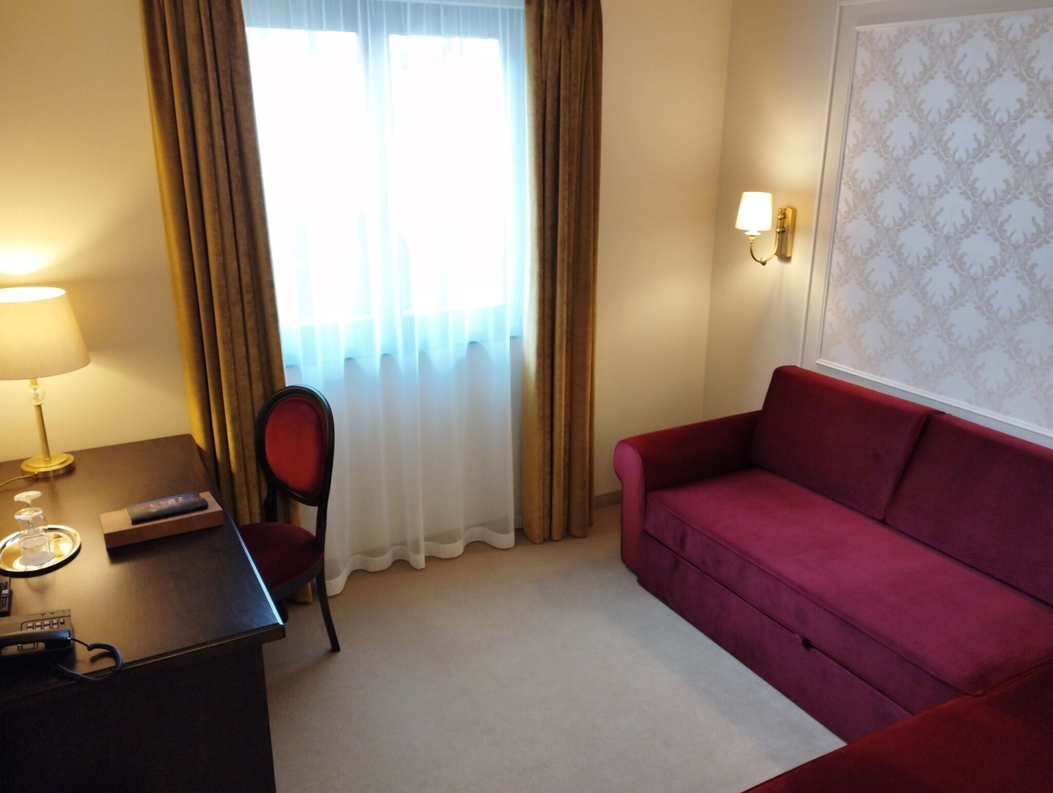 A hotel room with a desk, chair, red sofa, and a window with curtains.