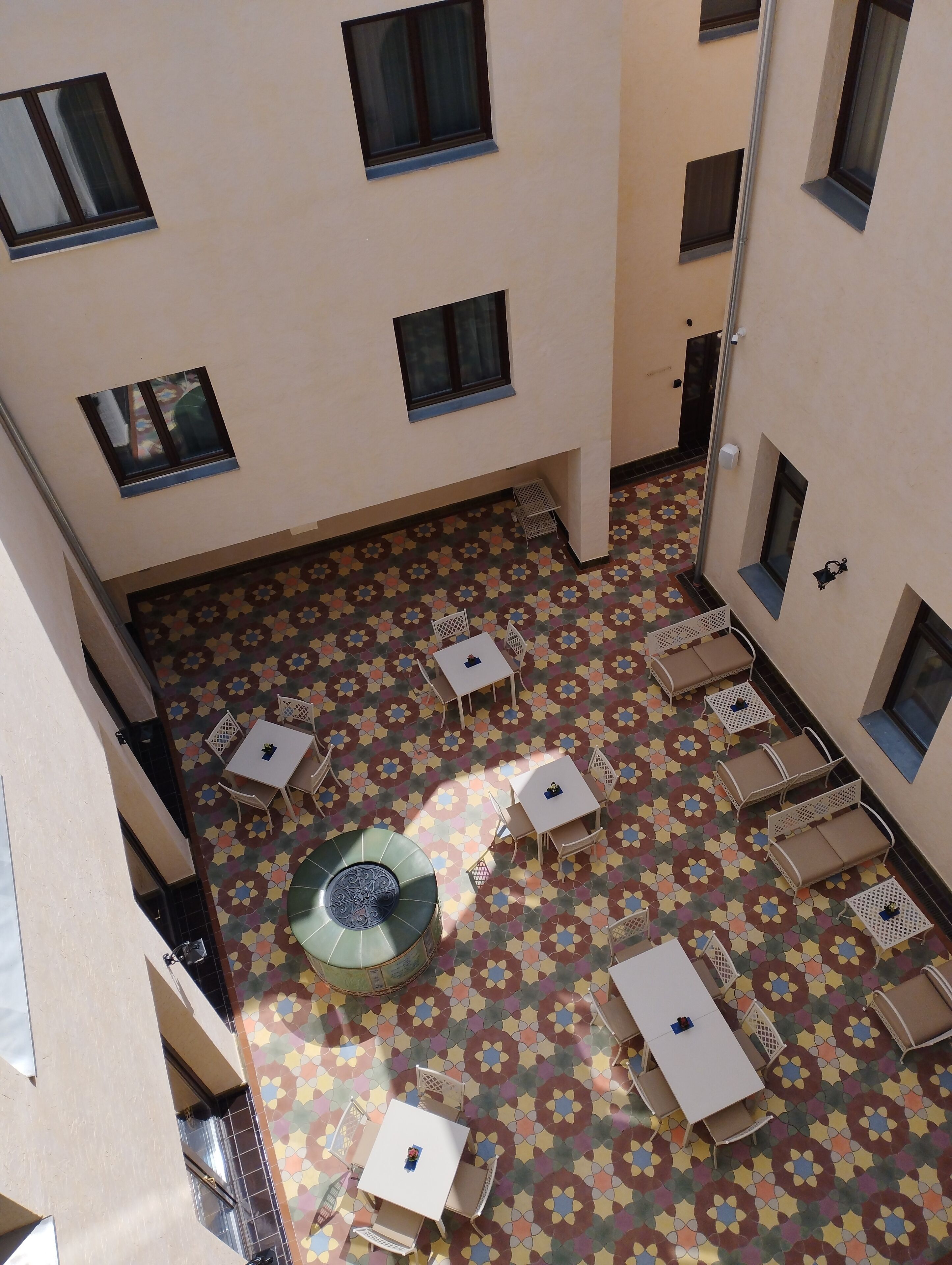 An outdoor courtyard with multiple tables and chairs, a decorative fountain, and patterned flooring.