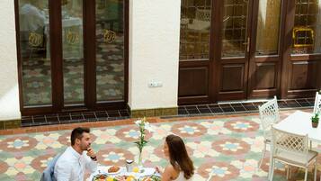 A dining area with a table set for two, a tiled floor, and a large arched window with a view of the interior.