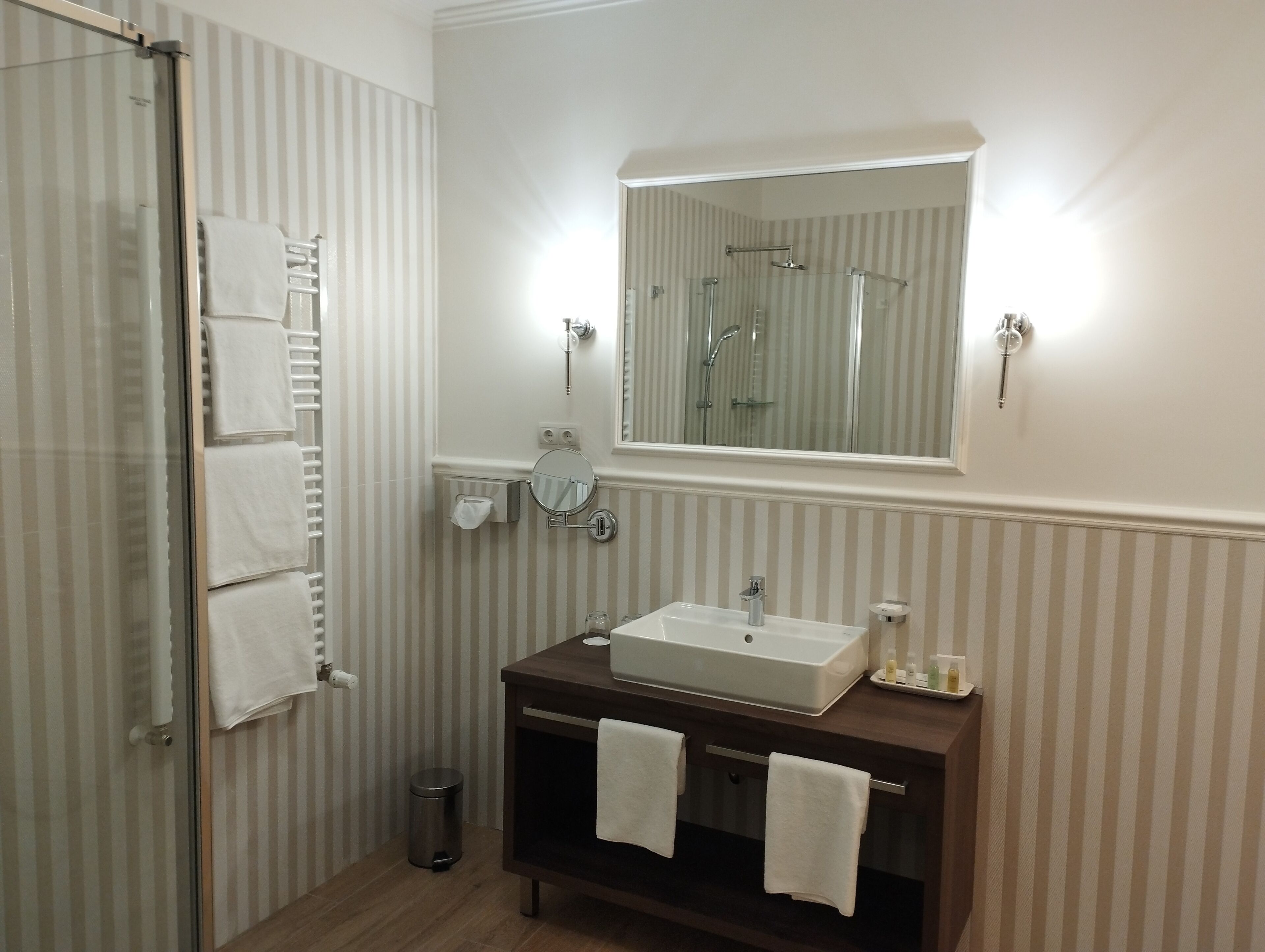 A bathroom with a shower, a sink, a mirror, and striped wallpaper.