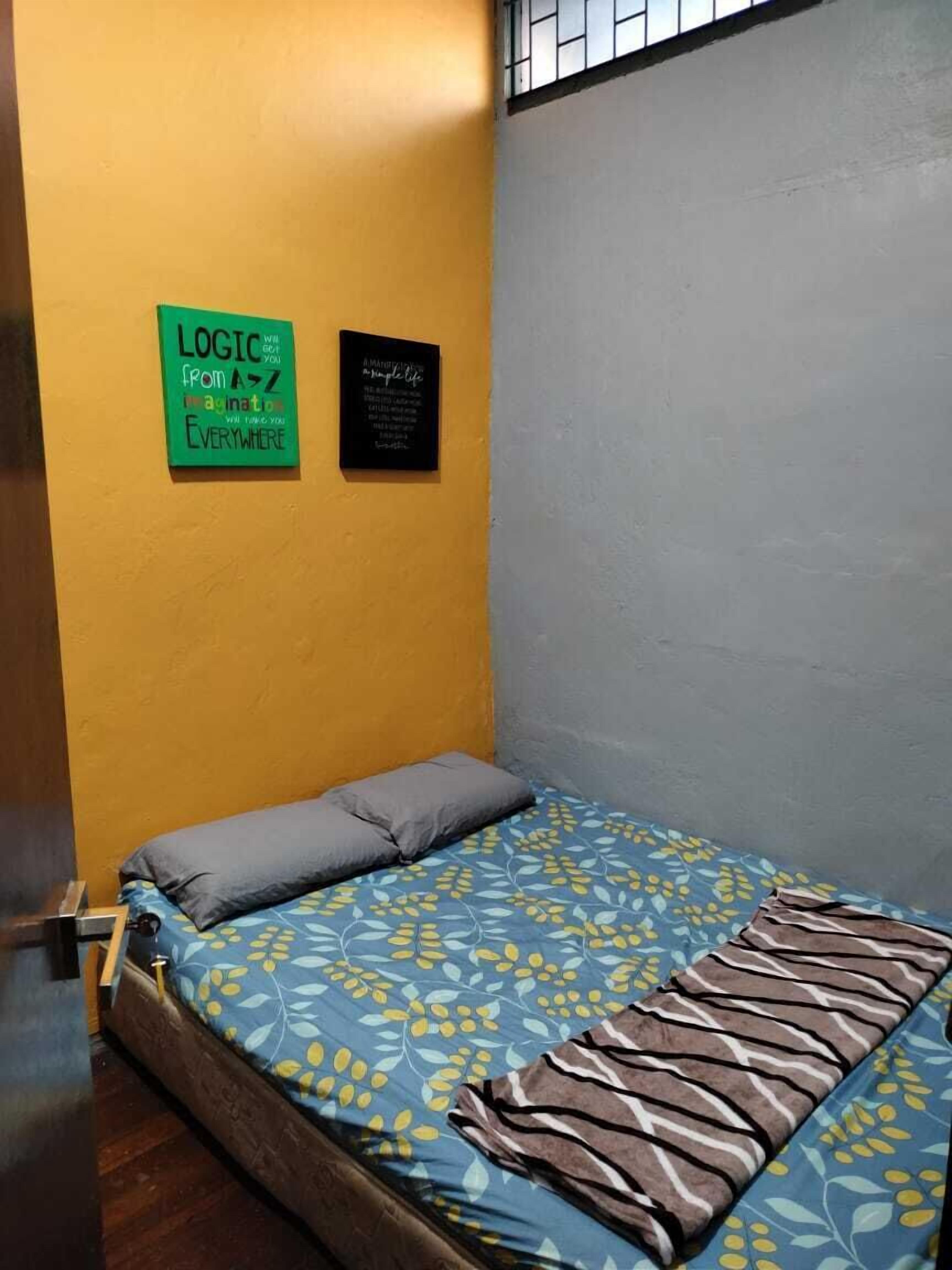 Una habitación pequeña con una cama individual, una pared amarilla y una pared gris. Hay dos letreros en la pared amarilla: uno verde con texto y otro negro con texto. La cama tiene una colcha de flores azules y amarillas y dos almohadas.