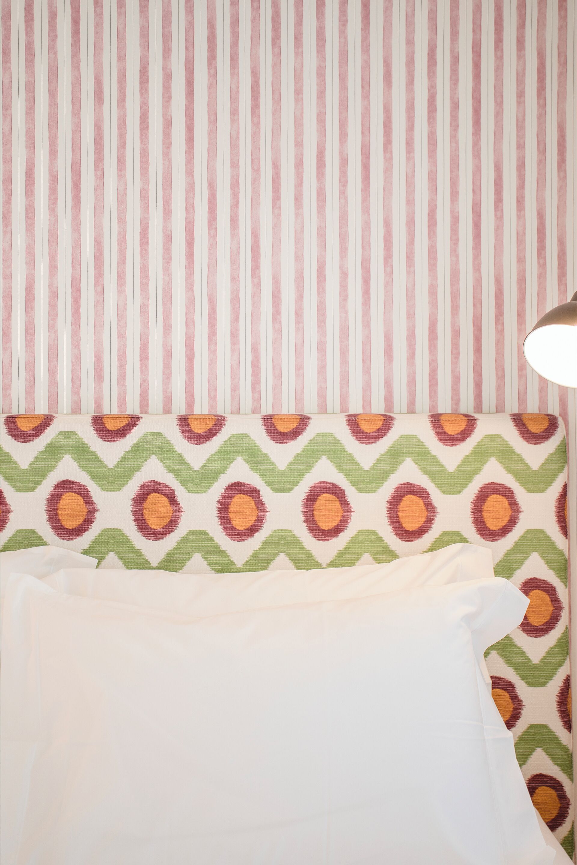 A bed with a white pillow and a patterned headboard against a striped wall.