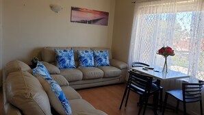 Een woonkamer met een beige bank, een eettafel met stoelen, een vaas met bloemen en een schilderij aan de muur.