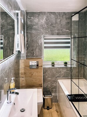 Un baño moderno con pared de mármol, tocador de madera, bañera y ventana con vista a un campo verde.