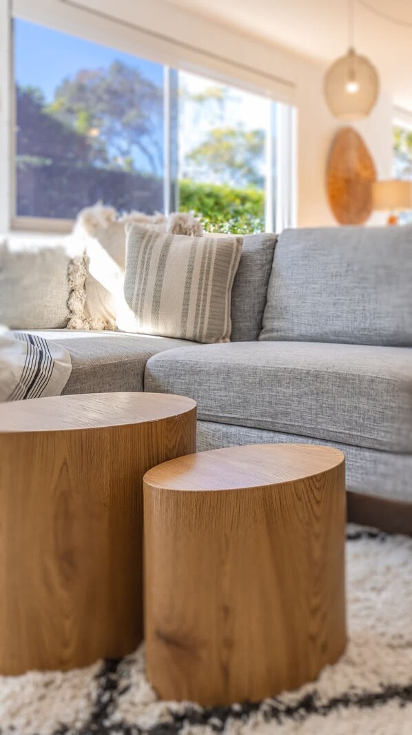 A living room with a grey sofa, two wooden side tables, and a large window with a view of greenery.