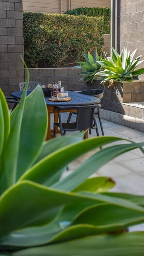 An outdoor seating area with a table and chairs, surrounded by potted plants and a hedge.