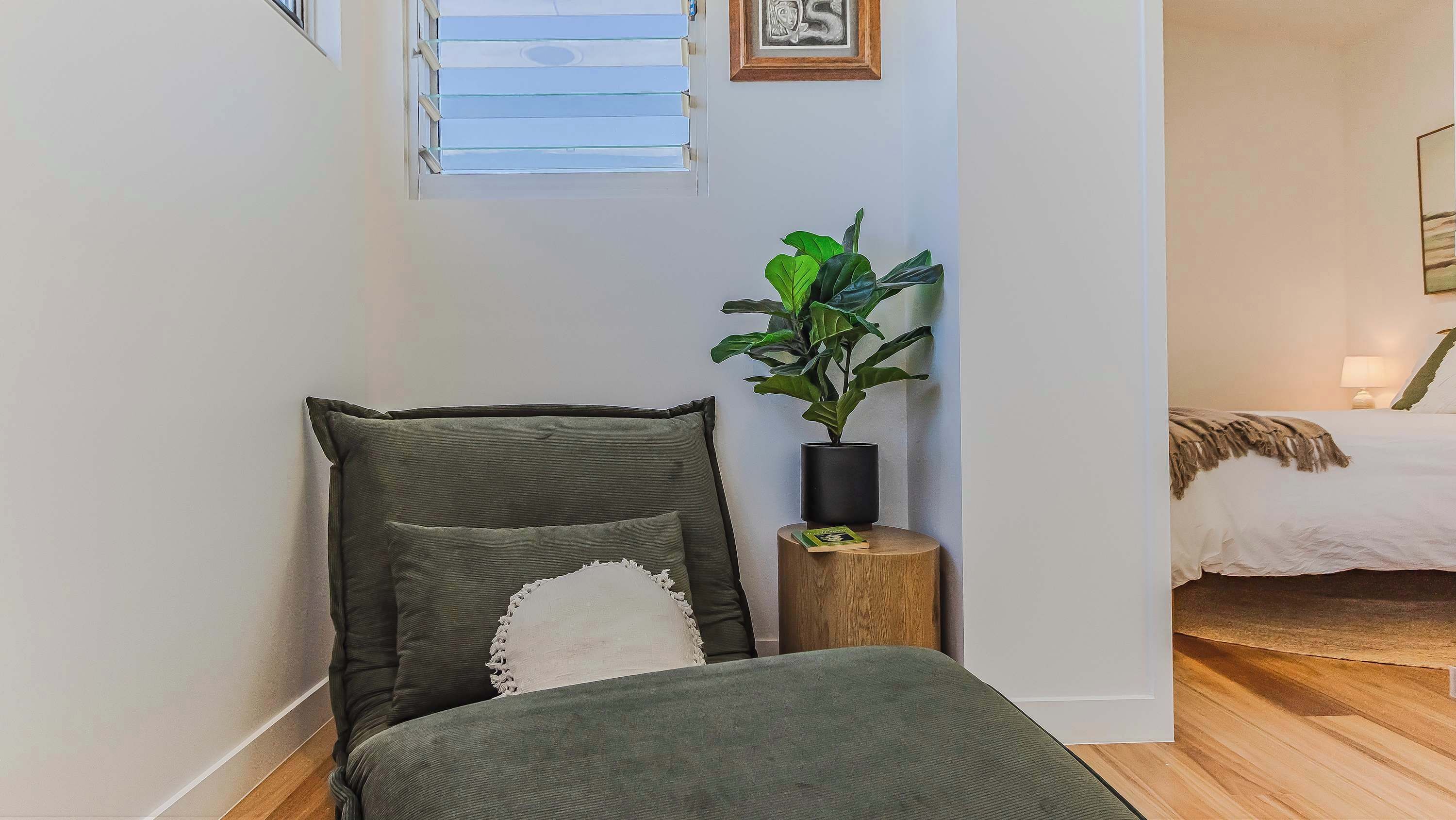 A corner of a room with a bed, a plant, and a window with blinds.