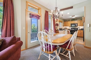 A dining area with a wooden table and white chairs, a chandelier, and a view of the kitchen and balcony. - 2 bedroom ski-in condo with hot tub, pool, and EV chargers (Killington)