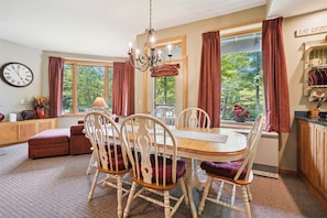 A dining area with a wooden table and white chairs, a chandelier, and a clock on the wall.