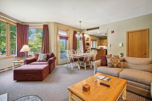 A spacious living room with a large window, a dining area, and a kitchen visible through an open door.