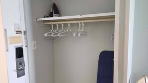 Habitación de hotel con una puerta blanca, un perchero con colgadores, una tabla de planchar azul y un pequeño dispositivo electrónico en el suelo.