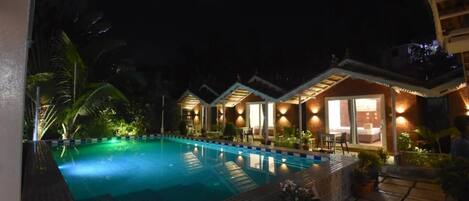 A nighttime view of a hotel with a swimming pool, well-lit rooms, and a tropical garden.