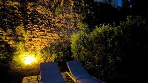 Un muro de piedra con una fuente de luz en la base, dos sillones de descanso y un camino de piedra.