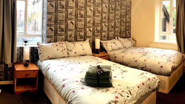 A bedroom with two beds, a wall of framed bird illustrations, a bedside table with a lamp, and a window with curtains. - Charming Three Bedroom 5 Double Bed Apt in London (London)