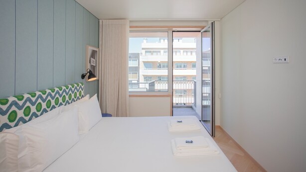 A hotel room with a bed, a window with a view of buildings, a door, and a wall-mounted light fixture.
