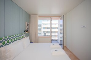 A hotel room with a bed, a window with a view of buildings, a door, and a wall-mounted light fixture. - Allegro Boutique Apartment C (Porto)