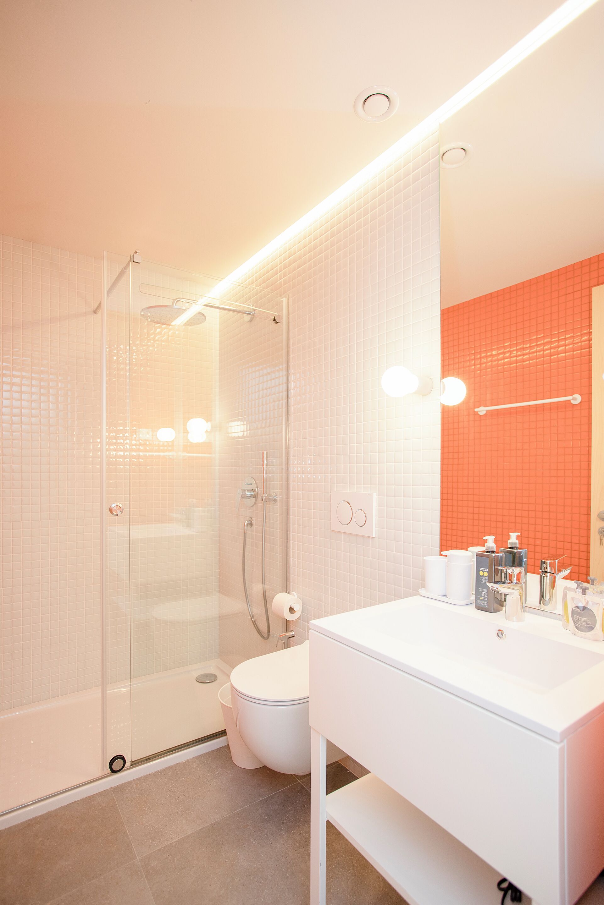 A modern bathroom with a glass-enclosed shower, a white sink with toiletries, and a mirror with an orange accent wall.