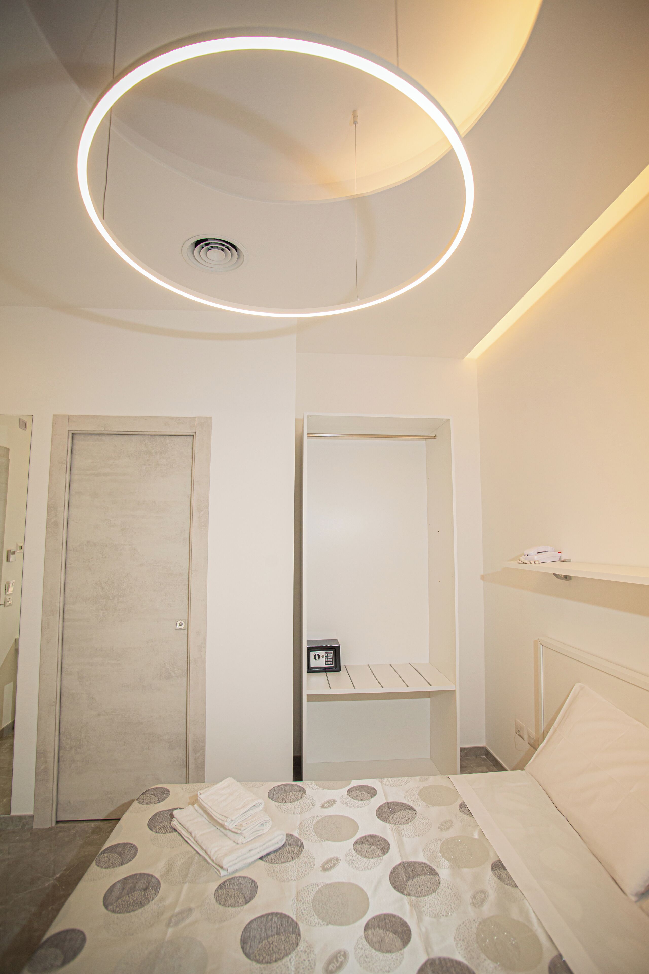 Habitación de hotel moderna con cama, lámpara de techo circular, armario y mesita de noche con teléfono.