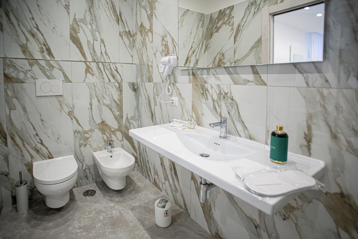 A modern bathroom with a marble backsplash, a white sink, a bidet, and a hairdryer mounted on the wall.
