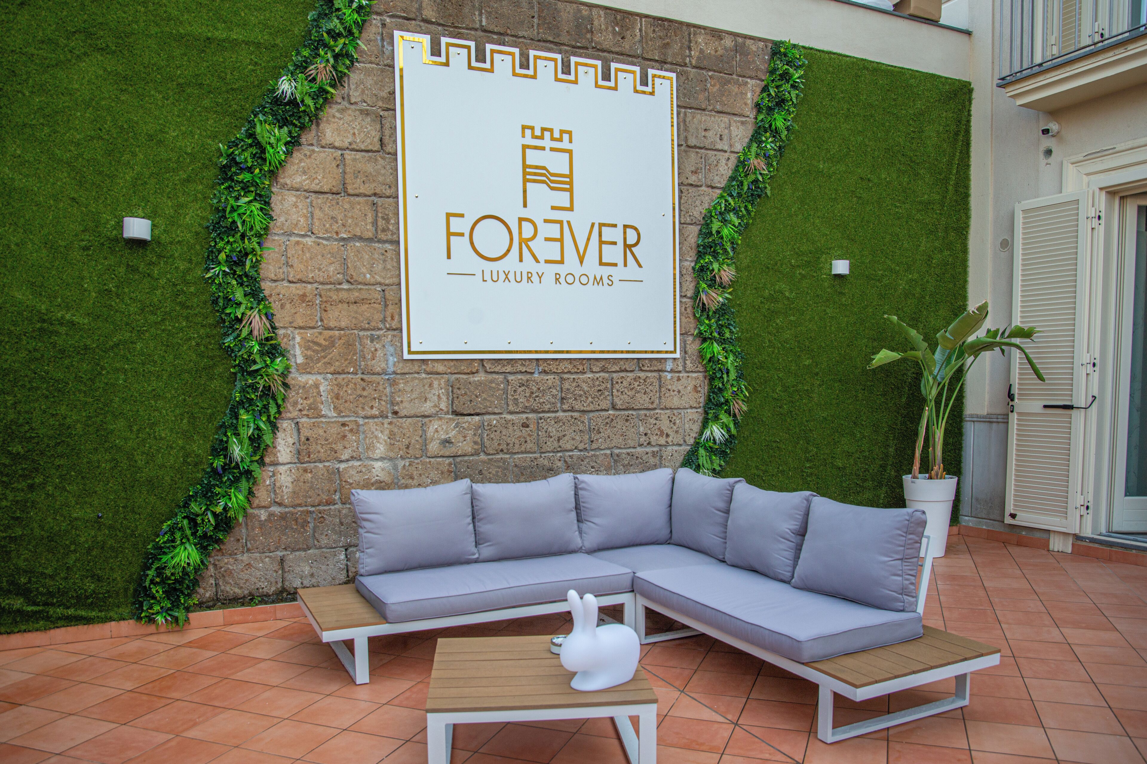 Uma área de estar moderna ao ar livre, com um sofá cinza e uma mesa de madeira, uma placa com 'FOREVER LUXURY ROOMS' e uma parede verde com plantas.