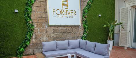 Uma área de estar moderna ao ar livre, com um sofá cinza e uma mesa de madeira, uma placa com 'FOREVER LUXURY ROOMS' e uma parede verde com plantas.