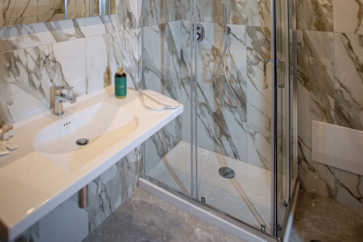 Un baño moderno con pared de mármol, cabina de ducha de vidrio y lavamanos blanco.