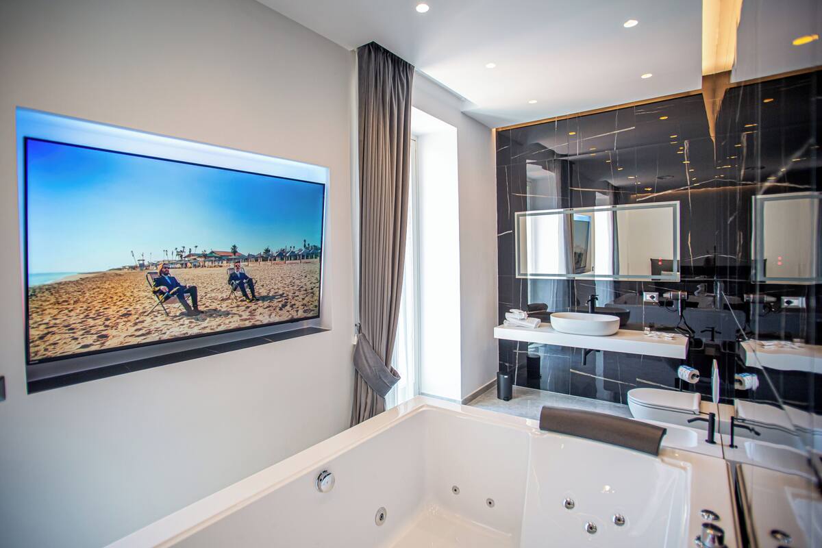 Un baño moderno con una bañera grande, un televisor empotrado mostrando una escena de playa y un tocador contemporáneo con un espejo amplio.
