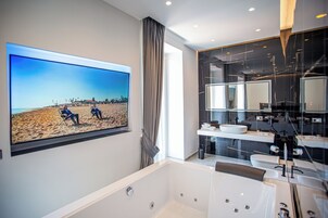 Un baño moderno con una bañera grande, un televisor empotrado mostrando una escena de playa y un tocador contemporáneo con un espejo amplio.