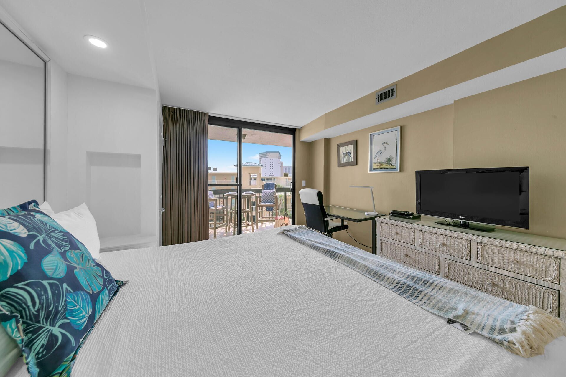 A hotel room with a large bed, a desk with a TV, and a balcony with a view of buildings.