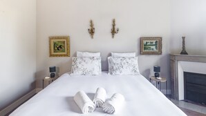 Una cama bien hecha con sábanas blancas y dos almohadas, una mesita de noche con lámpara y una chimenea al fondo.