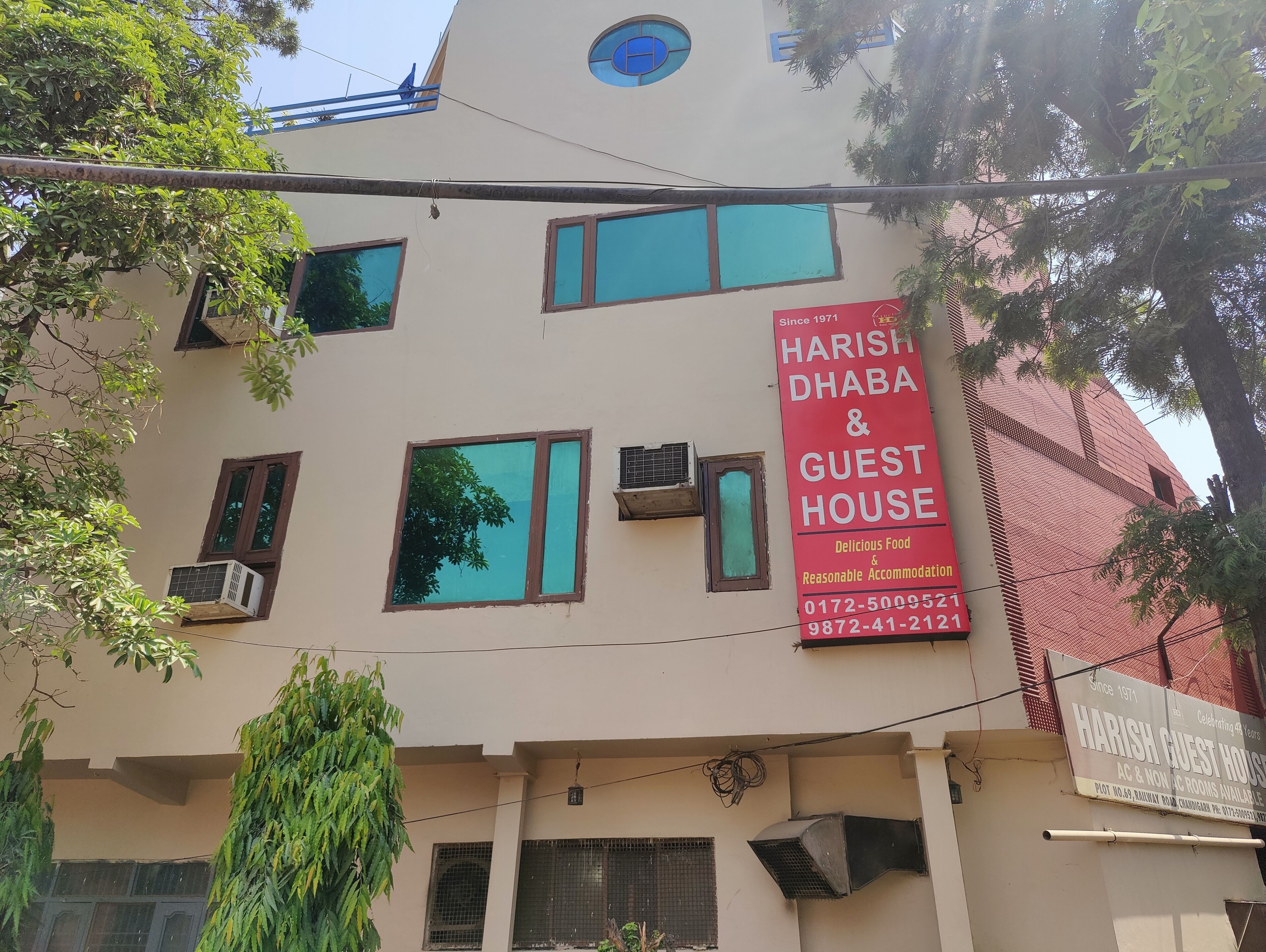 A building with multiple windows and air conditioning units, a prominent red sign advertising Harish Dhaba & Guest House, and a tree in the foreground.