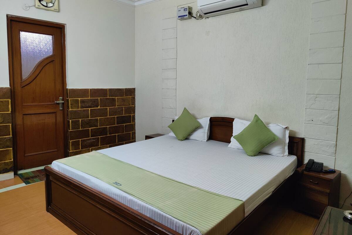 A neatly made bed with a green bedspread and pillows, a wooden door with a glass panel, and an air conditioning unit mounted on the wall.