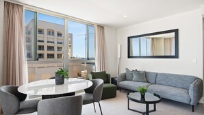 A modern living room with a round glass table, grey sofa, and a large window offering a view of a building. - Belle Escapes - Beachfront Balcony Glenelg (Glenelg)